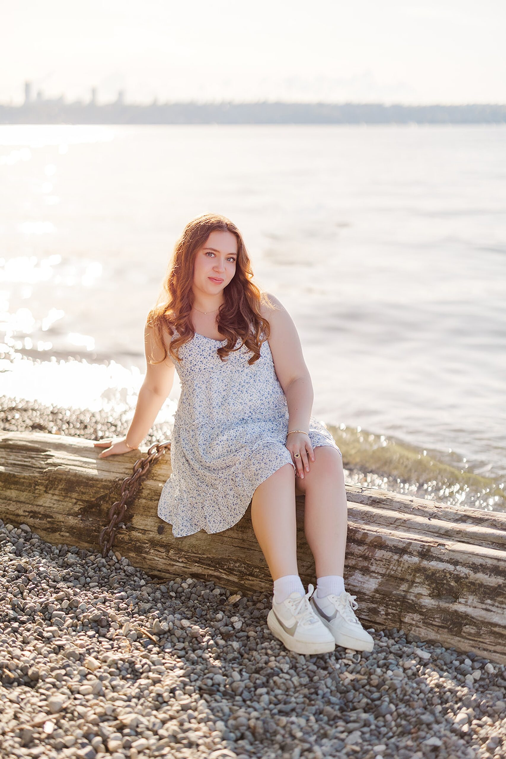 A high school senior in a white dress and sneakers sits on a log on a beach at sunset