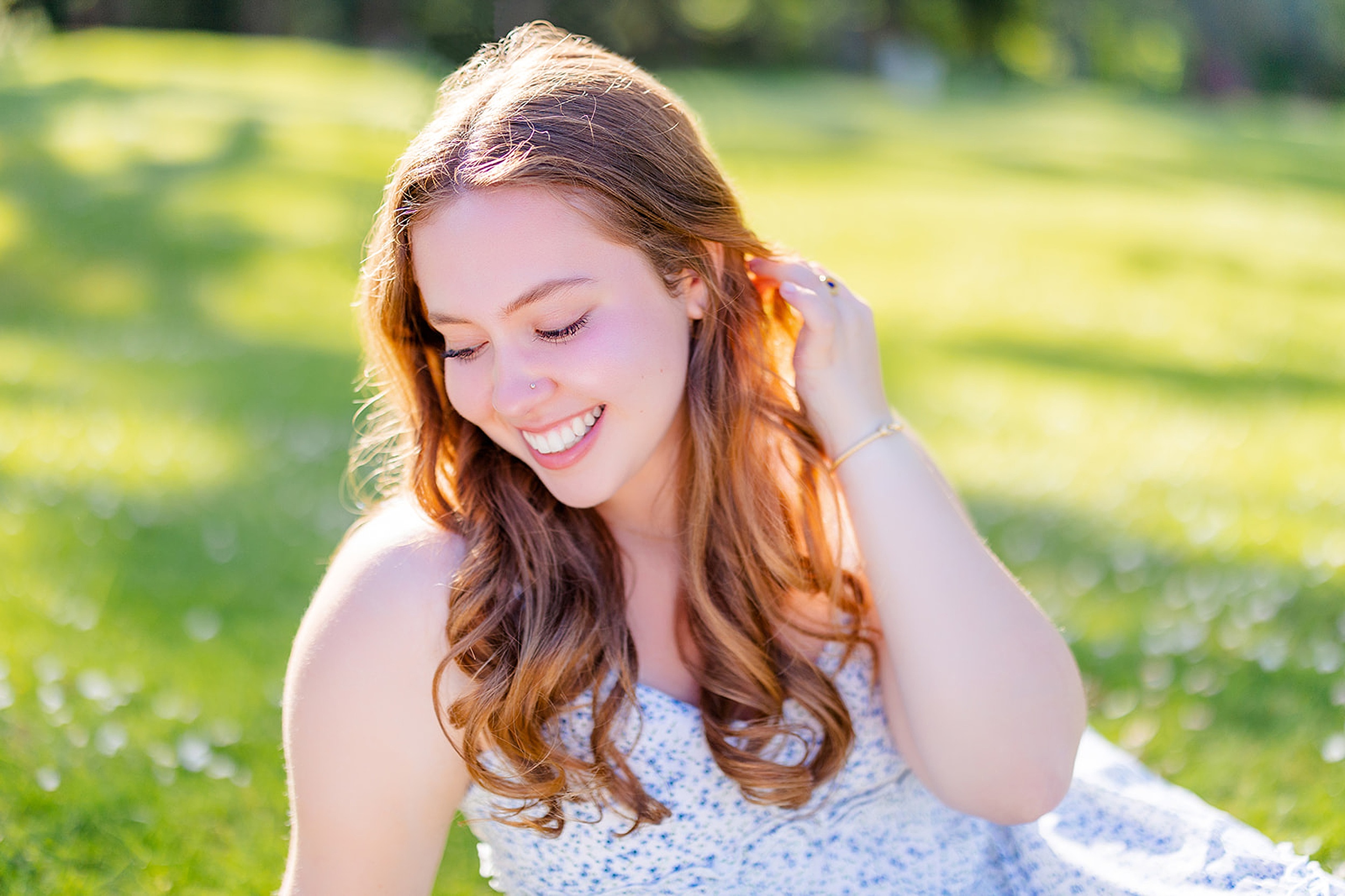 A teenage high school senior in a blue dress smiles down her shoulder while sitting in a green lawn at sunset before visiting colleges near issaquah wa