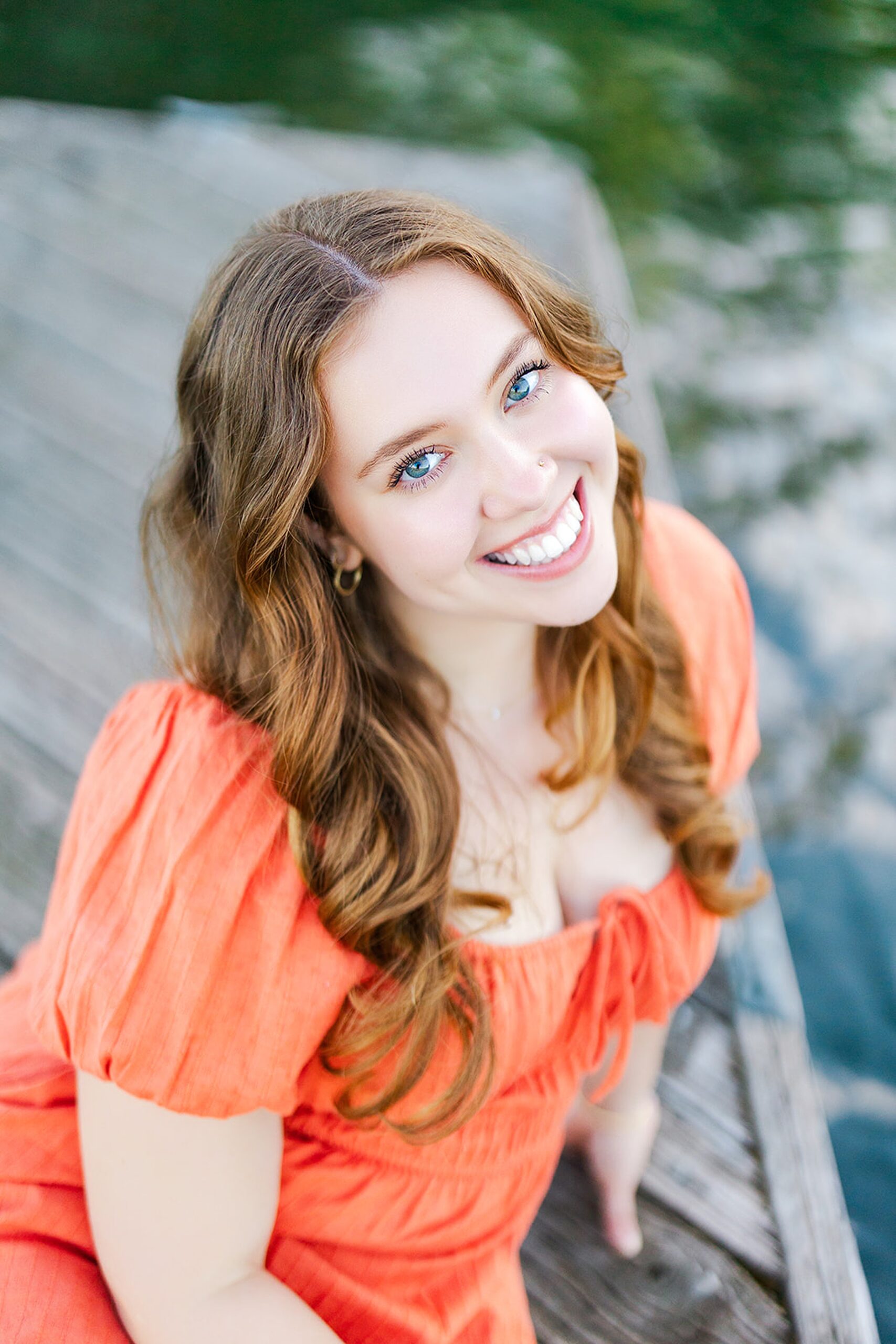 A red hair high school senior sits on a wooden dock smiling in an orange dress before visiting colleges near issaquah wa