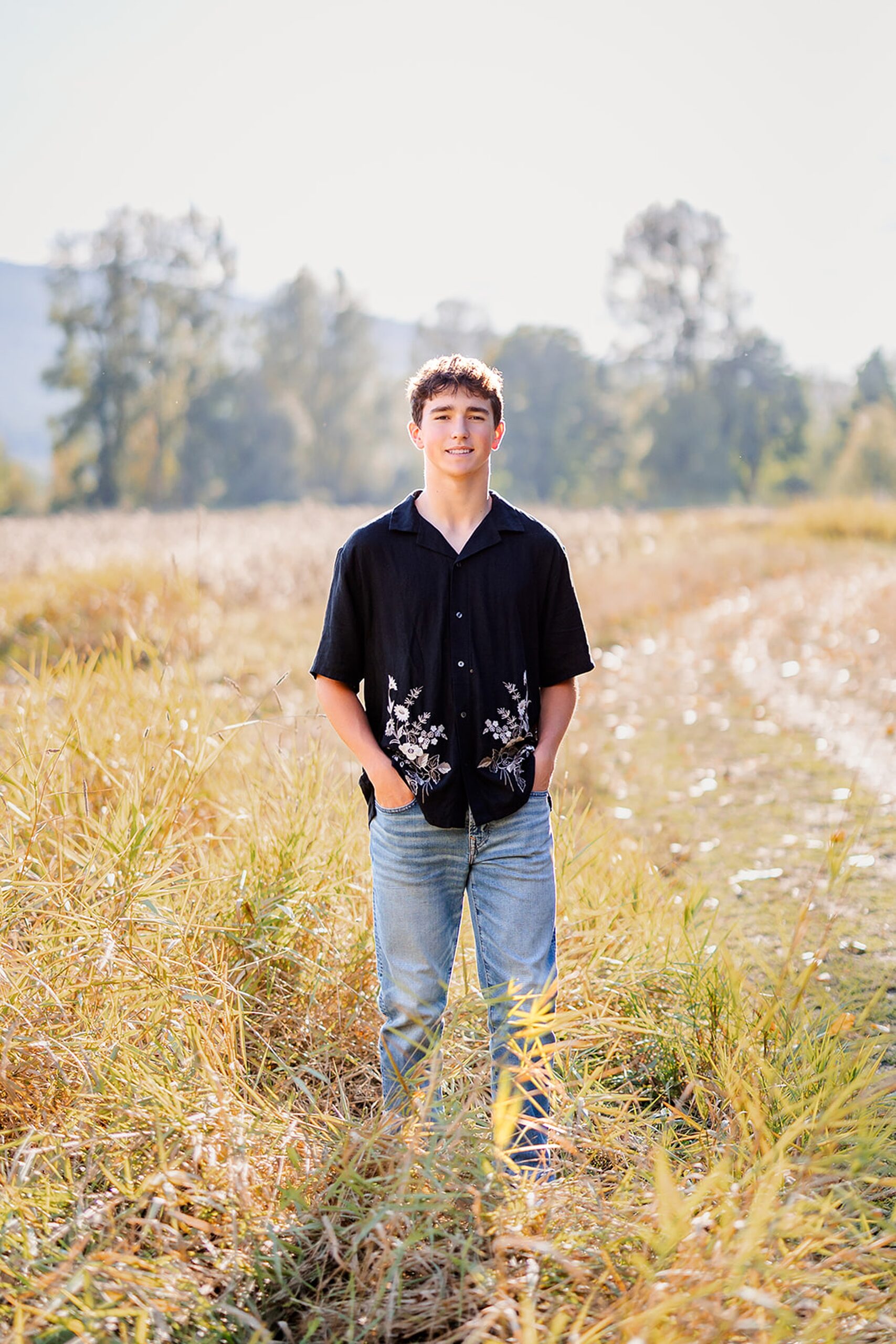 A teenage boy stands with hands in pockets in a black button down shirt in a park trail at sunset