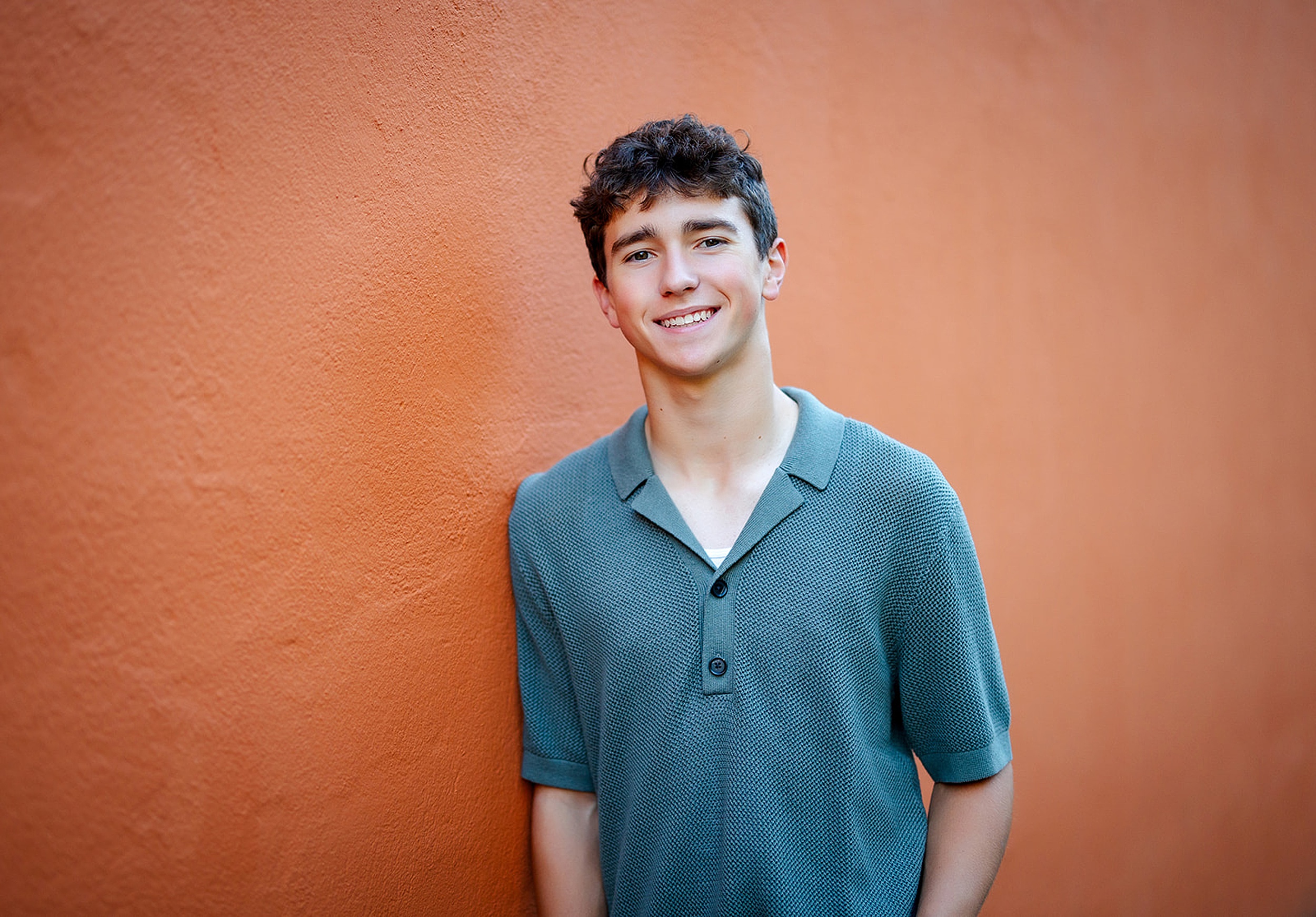 A smiling high school senior stands against and orange wall in a polo shirt before visiting Colleges Near Kirkland, WA