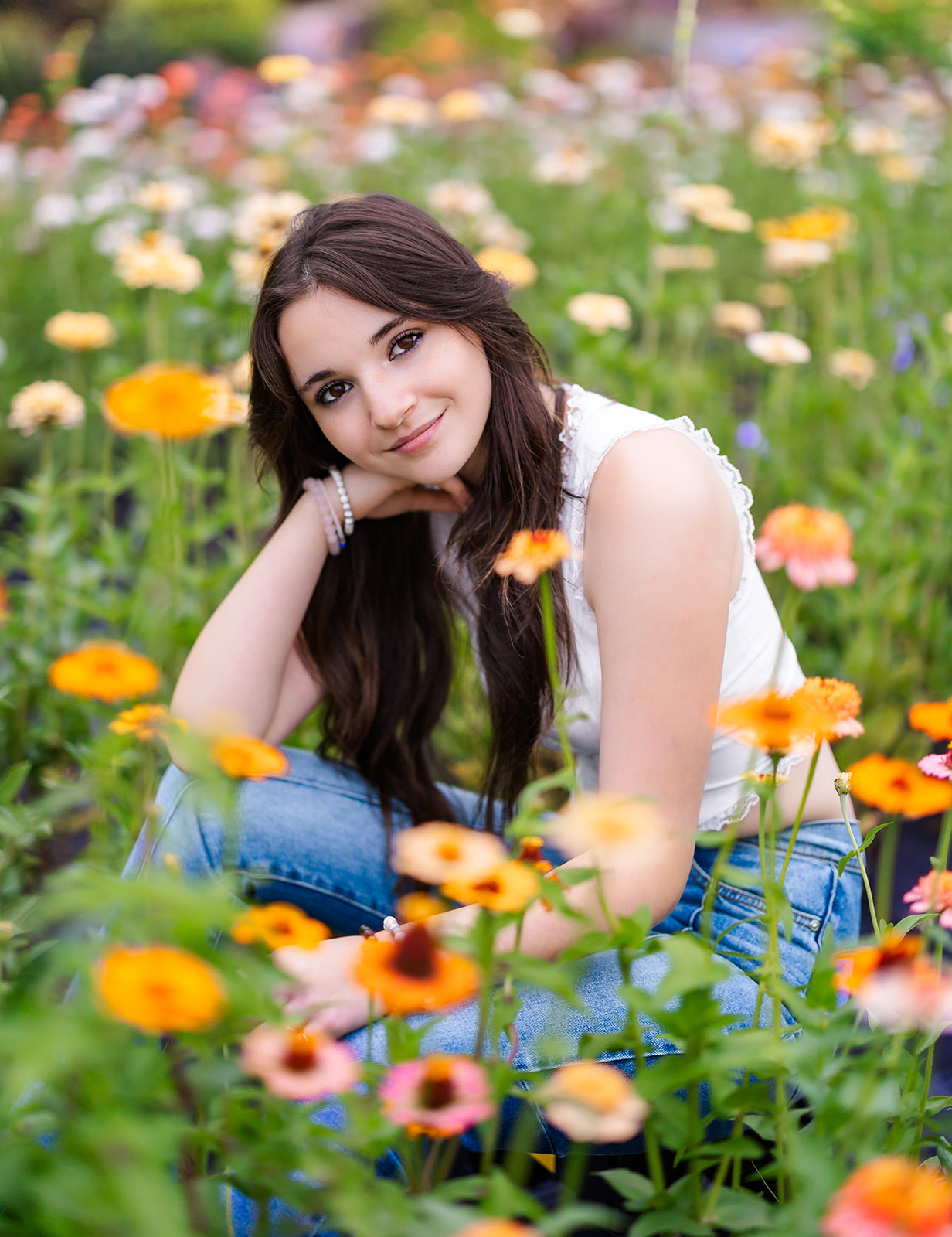 A high school senior with dark hair sits in a lush flower garden resting on her knee