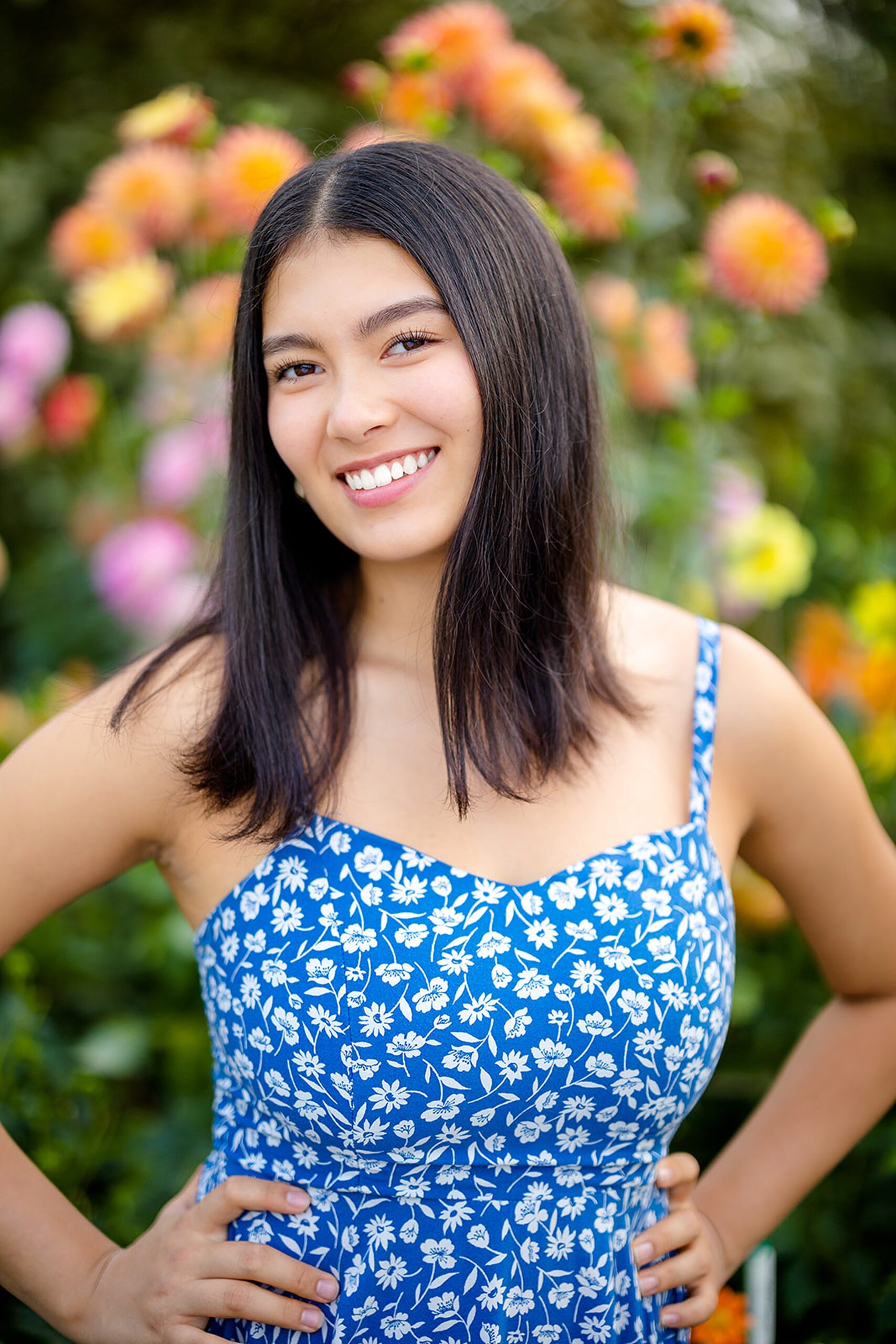 A smiling high school senior stands in a flowering garden with hands on her hips in a blue floral print dress from one of the Issaquah Boutiques