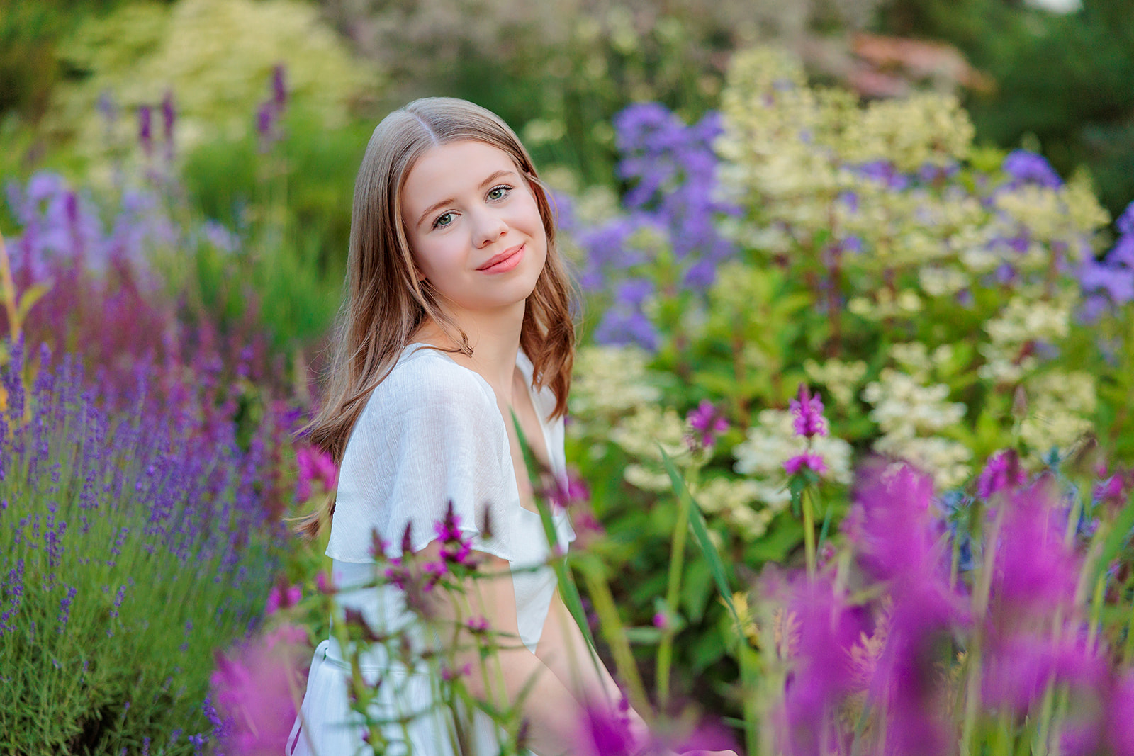 A teenage girl with brown hair sits in the middle of a colorful flower garden