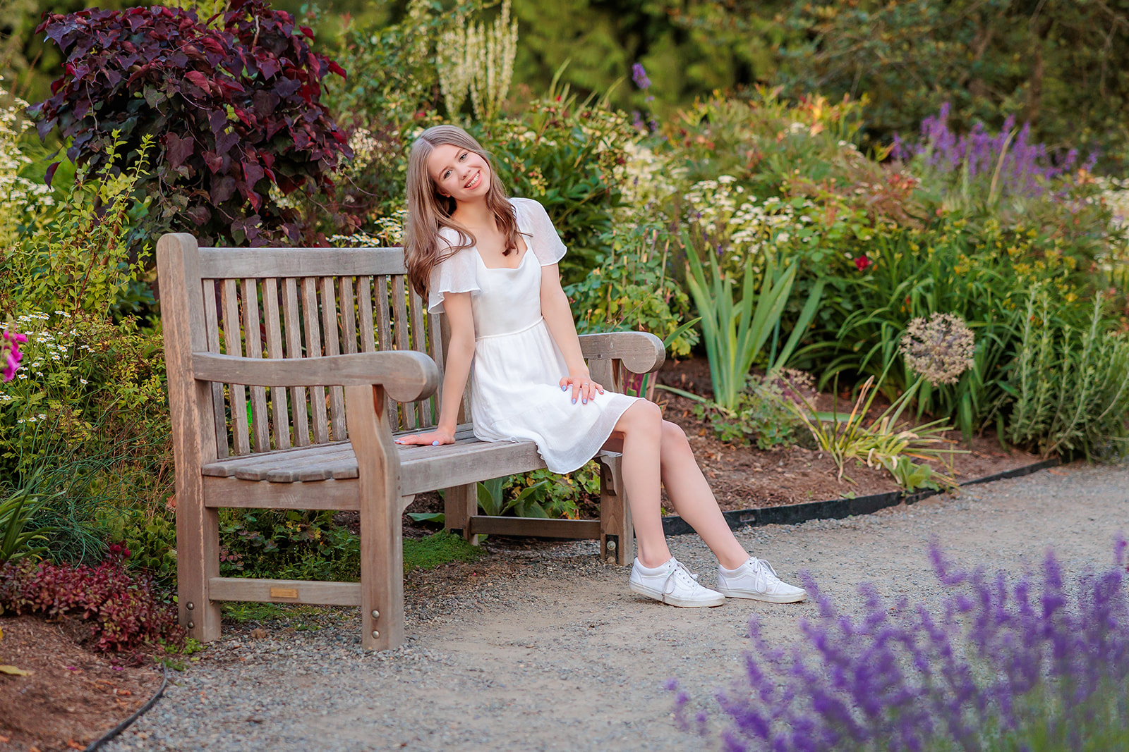 A teenager in a white dress sits on a wooden garden bench on a gravel path before visiting Issaquah Florists