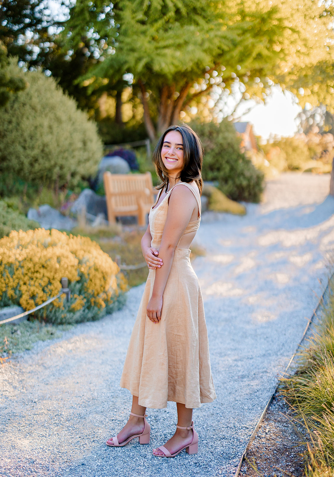 A dark hair high school senior walks in a garden trail at sunset in a tan dress