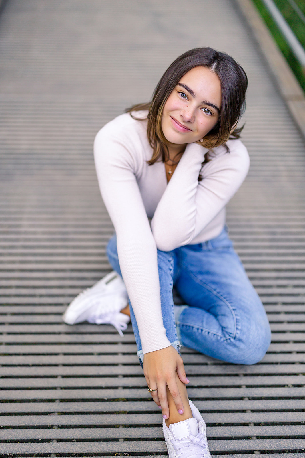 A dark hair high school senior in a white shirt and jeans sits on a park bridge at sunset after visiting Issaquah Hair Salons