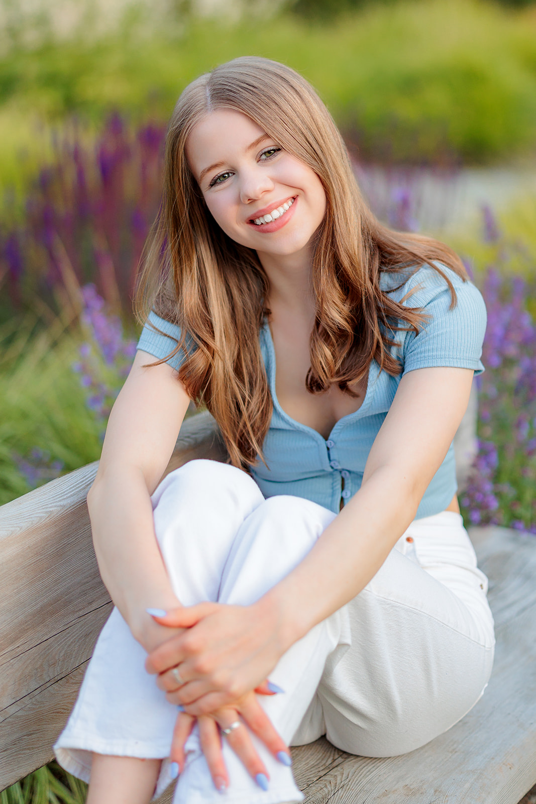 A high school senior in a blue top and matching nails sits across a wooden garden bench