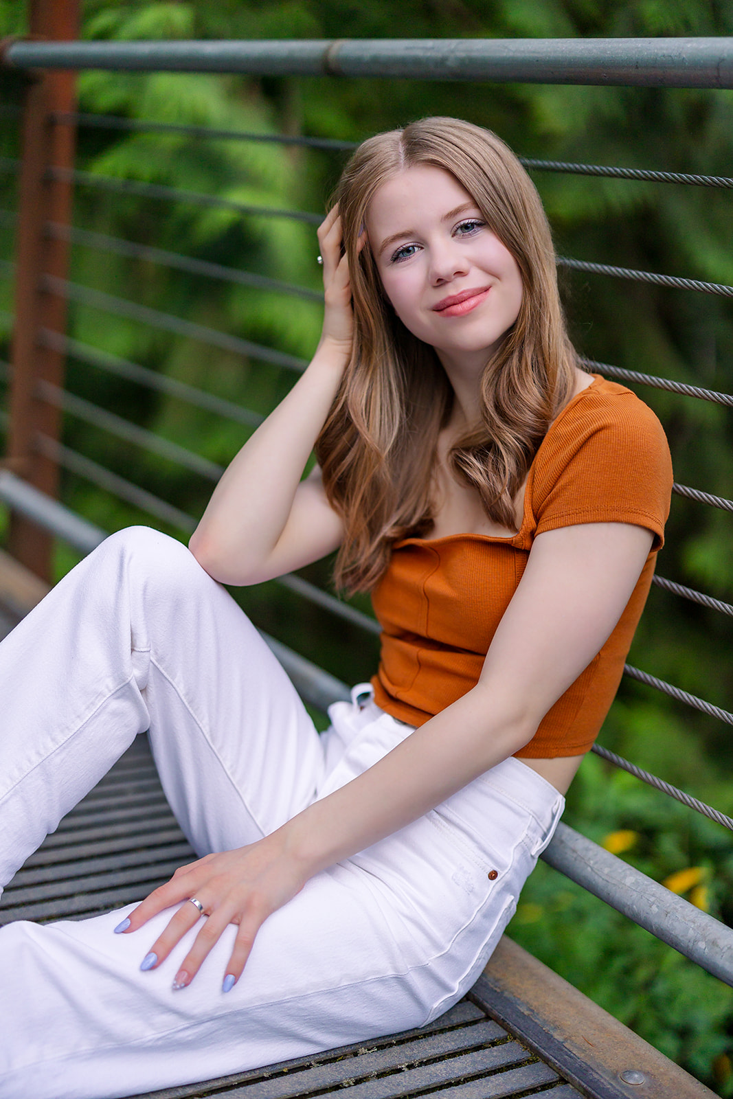 A high school senior in an orange top and white pants sits on a bridge leaning on the railing after visiting Issaquah Nail Salons