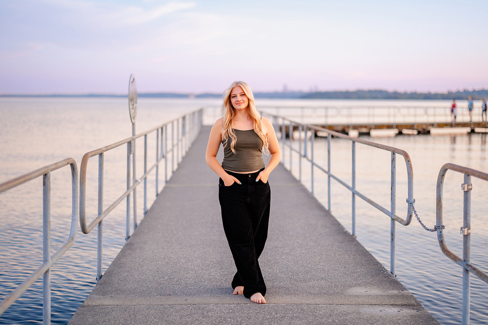 A high school senior in black pants walks barefoot on a long dock at sunset