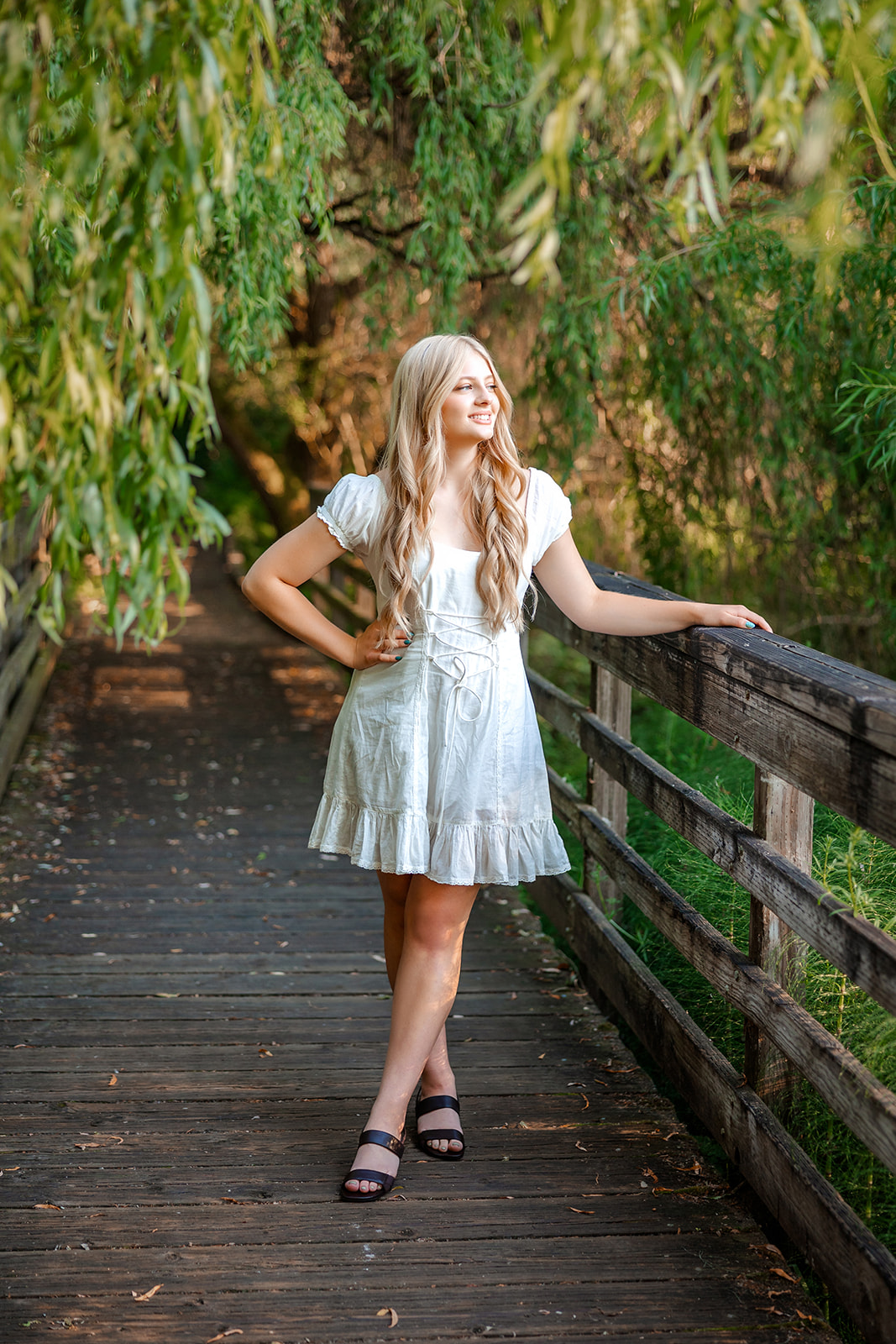 A blonde high school senior walks on a boardwalk in a white dress under a willow tree after visiting Issaquah Tanning Salons