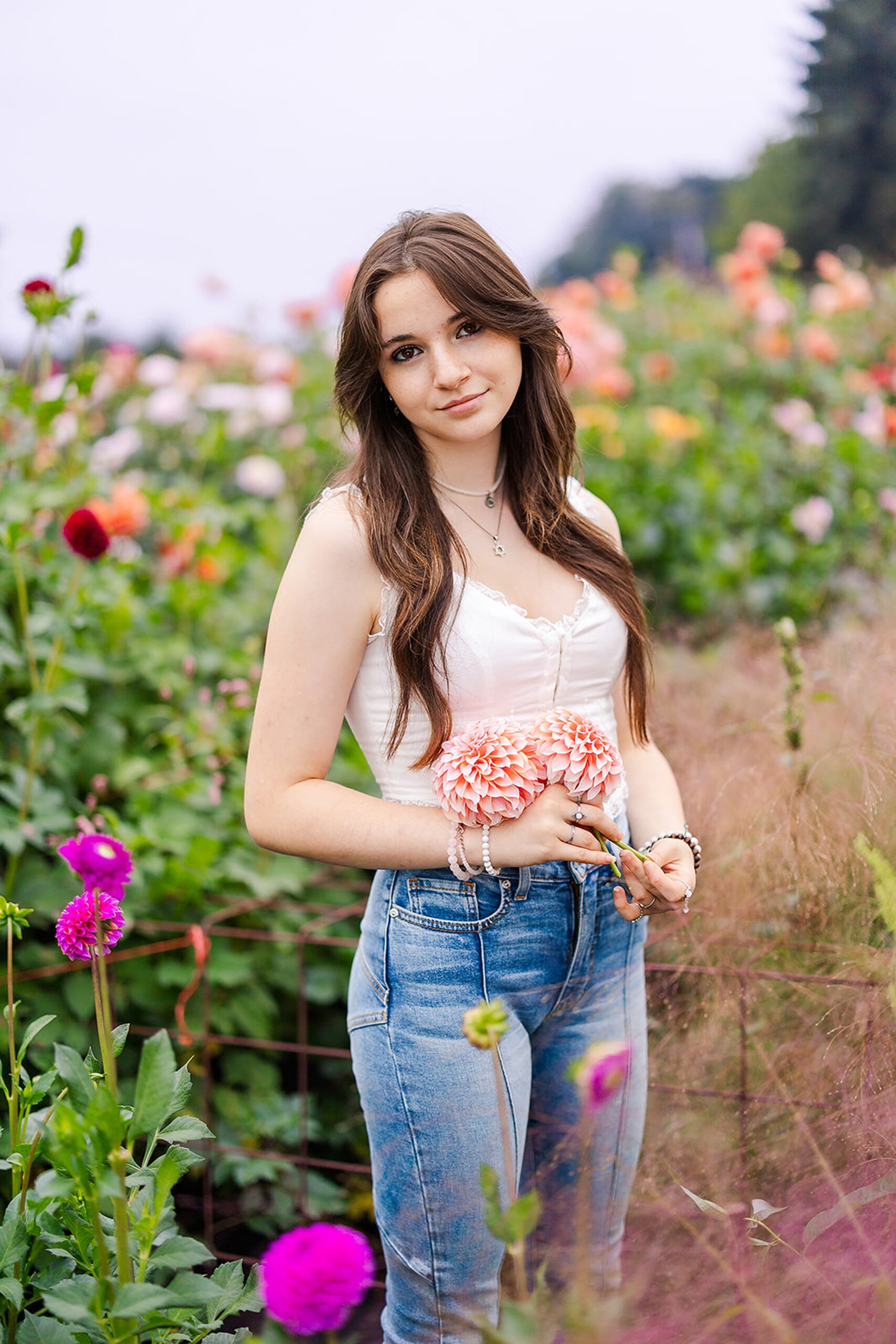 A high school senior walks through a flowering garden in jeans and a white top while holding pink flowers