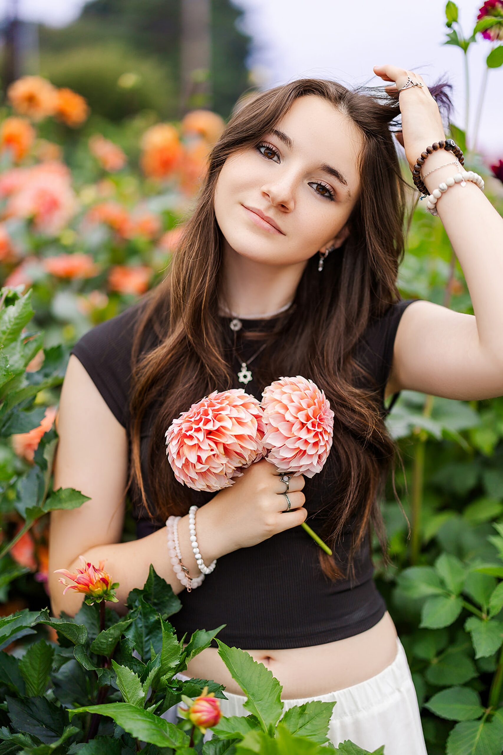 A high school senior holds some pink flowers while standing in a garden in a black top and white pants with a hand through her hair after shopping Kirkland Boutiques