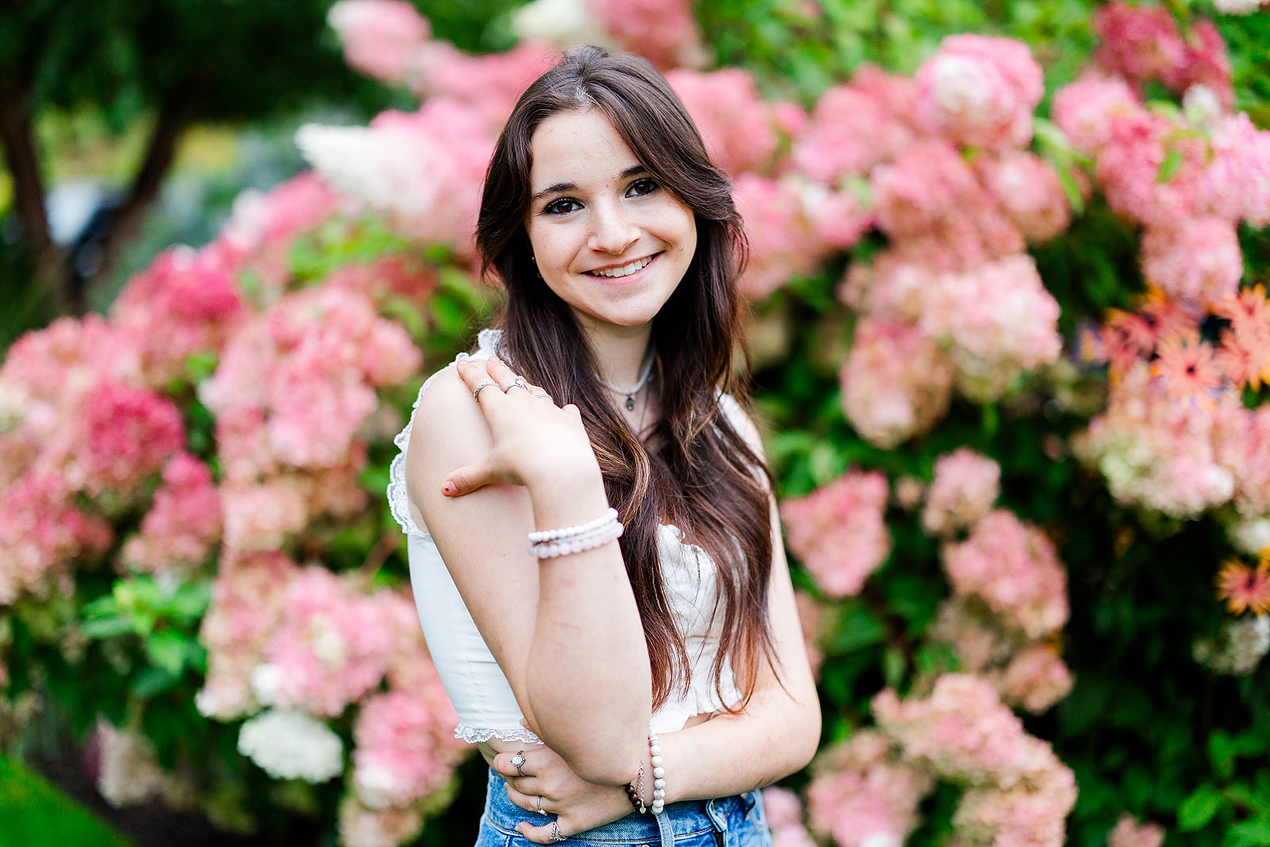 A high school senior with dark hair stands in a flowering garden in a white top and jeans after visiting Kirkland Boutiques