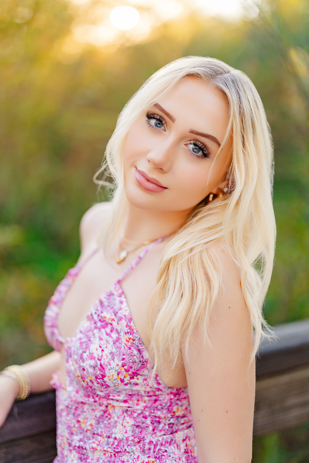 A blonde high school senior in a pink dress leans on a boardwalk railing at sunset