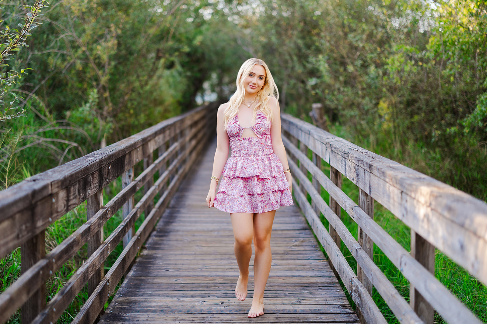 A high school senior with blonde hair walks on a wooden boardwalk at sunset barefoot in a pink dress after visiting Kirkland Hair Salons