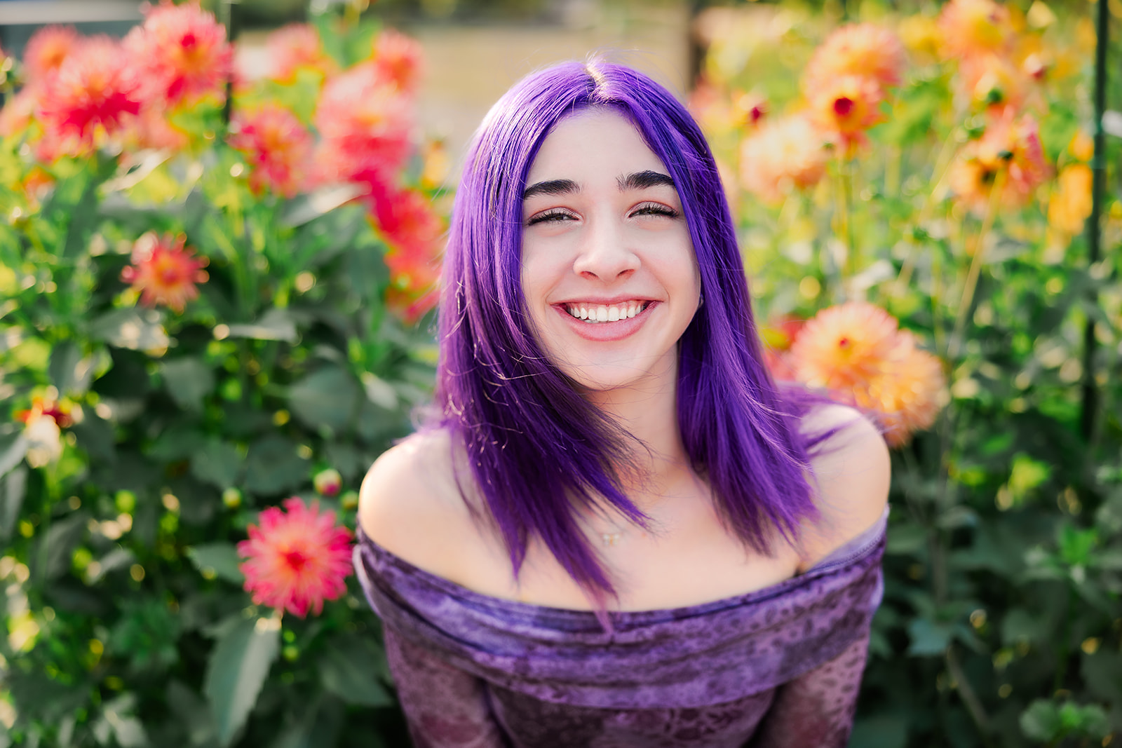 A happy high school senior with purple hair and a matching dress sits in a flower garden smiling