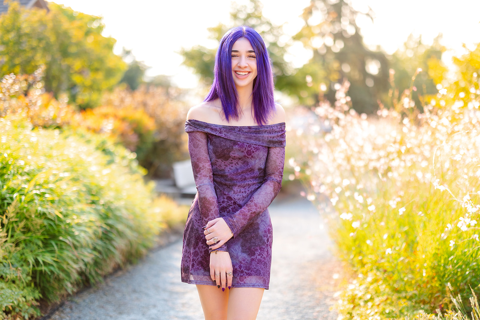 A high school senior in a purple dress with matching hair walks through a garden path at sunset after visiting Kirkland Nail Salons