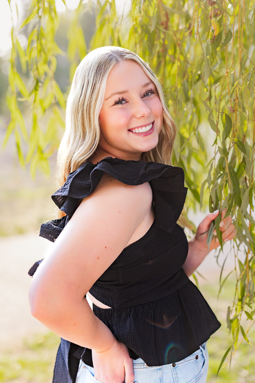 A blonde high school senior stands in a park in a black shirt smiling big while holding branches of a willow tree