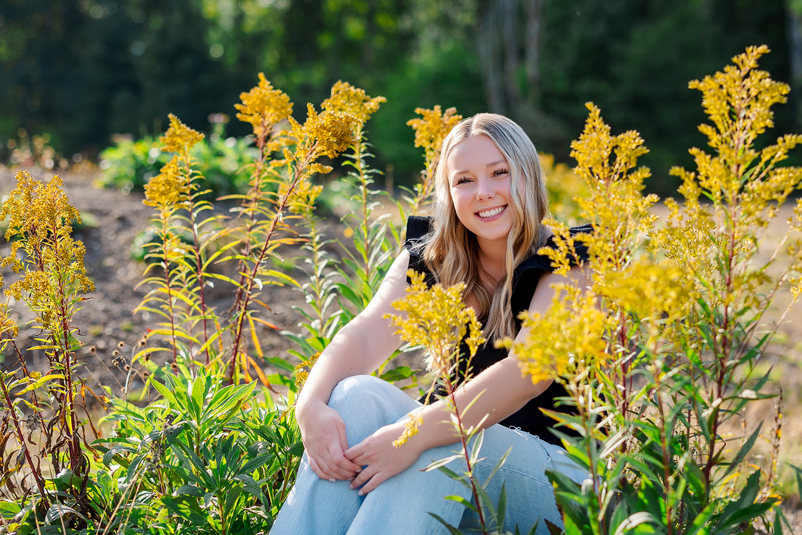 A high school senior wearing jeans and a black shirt sits among a patch of yellow flowers before visiting Kirkland Tanning Salons