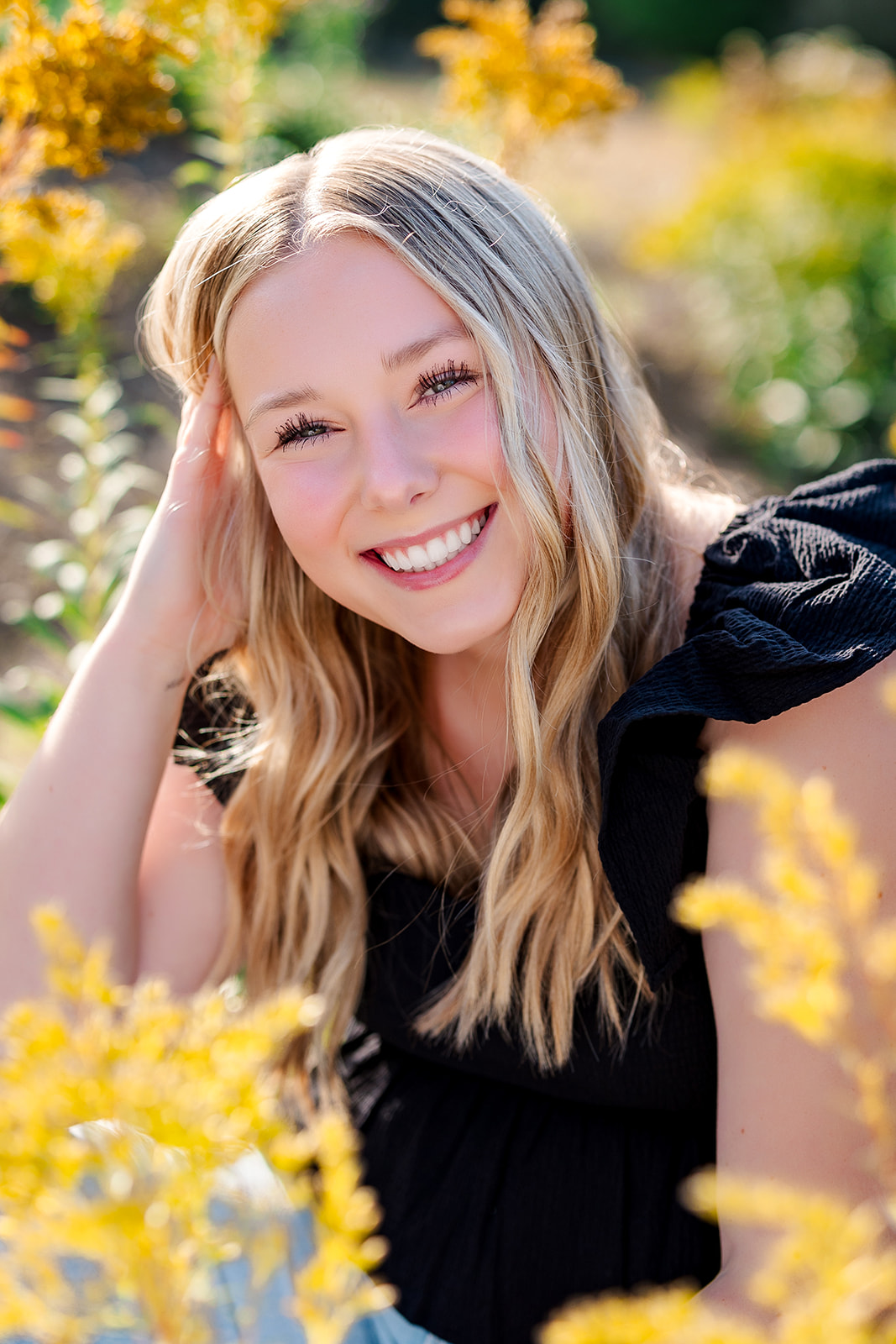 A high school senior smiles big while sitting in a field of wildflowers before visiting Kirkland Tanning Salons