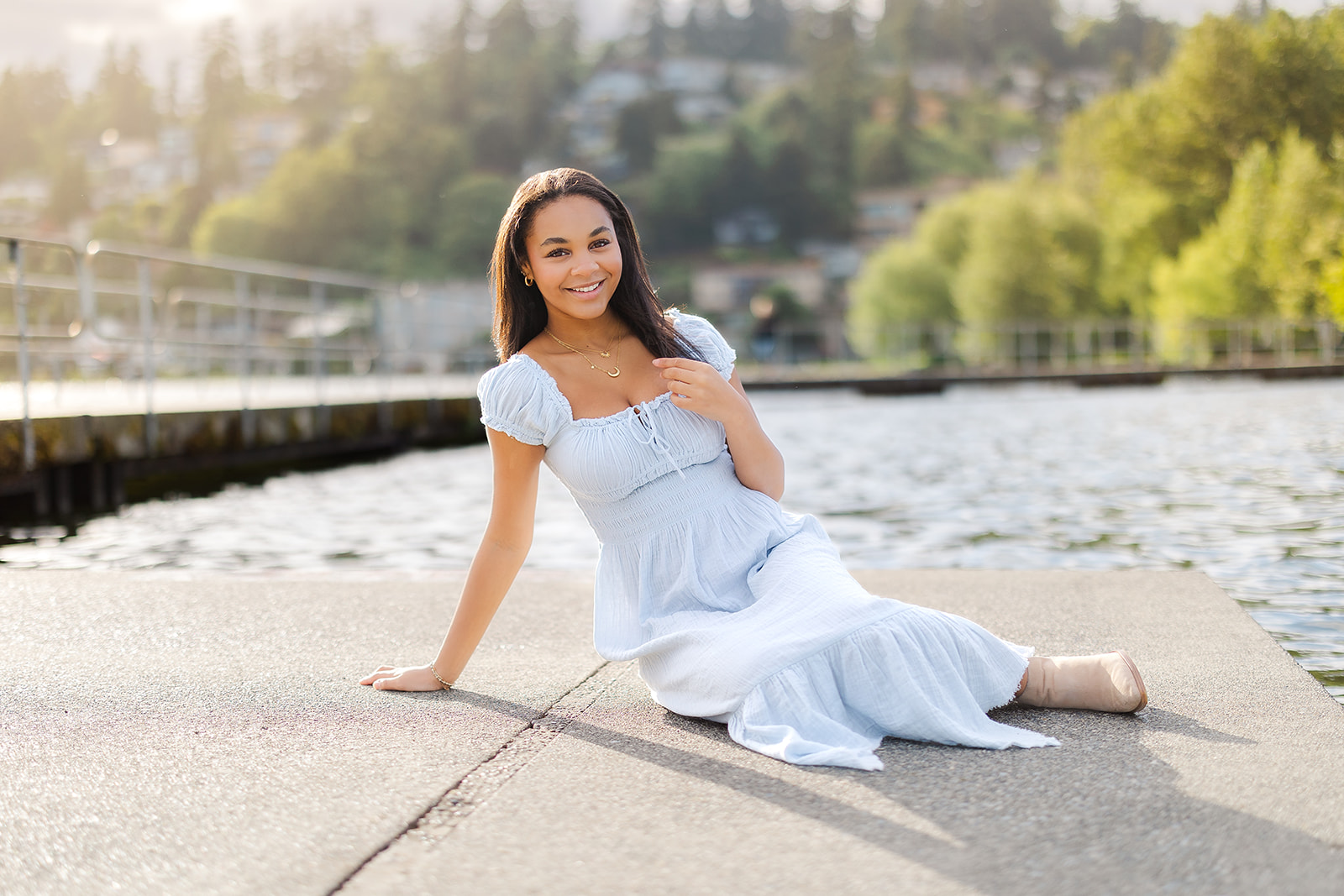 A high school senior in a baby blue dress sits on a cement dock at sunset