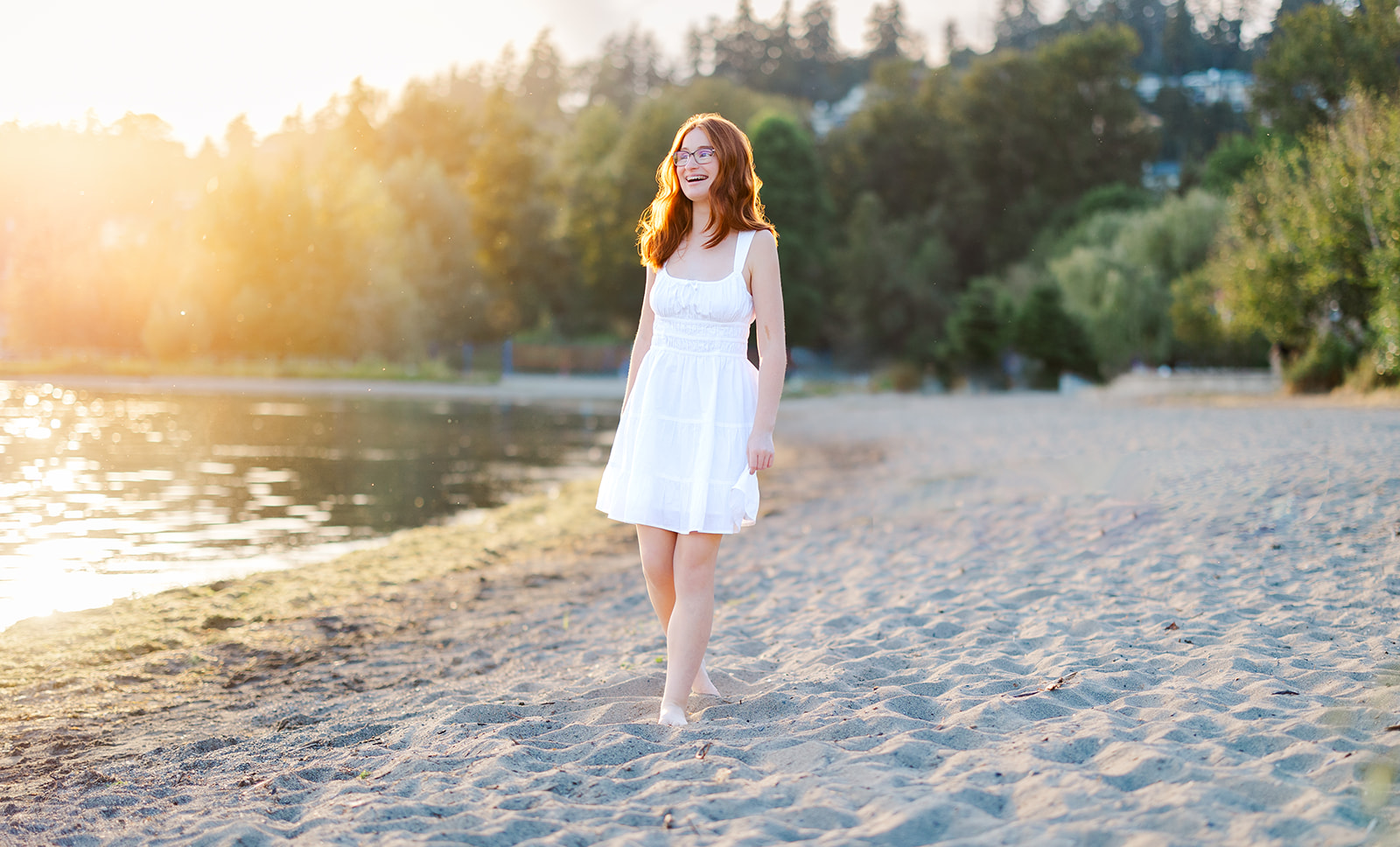 A teenage girl in a white dress laughs while walking up a beach at sunset