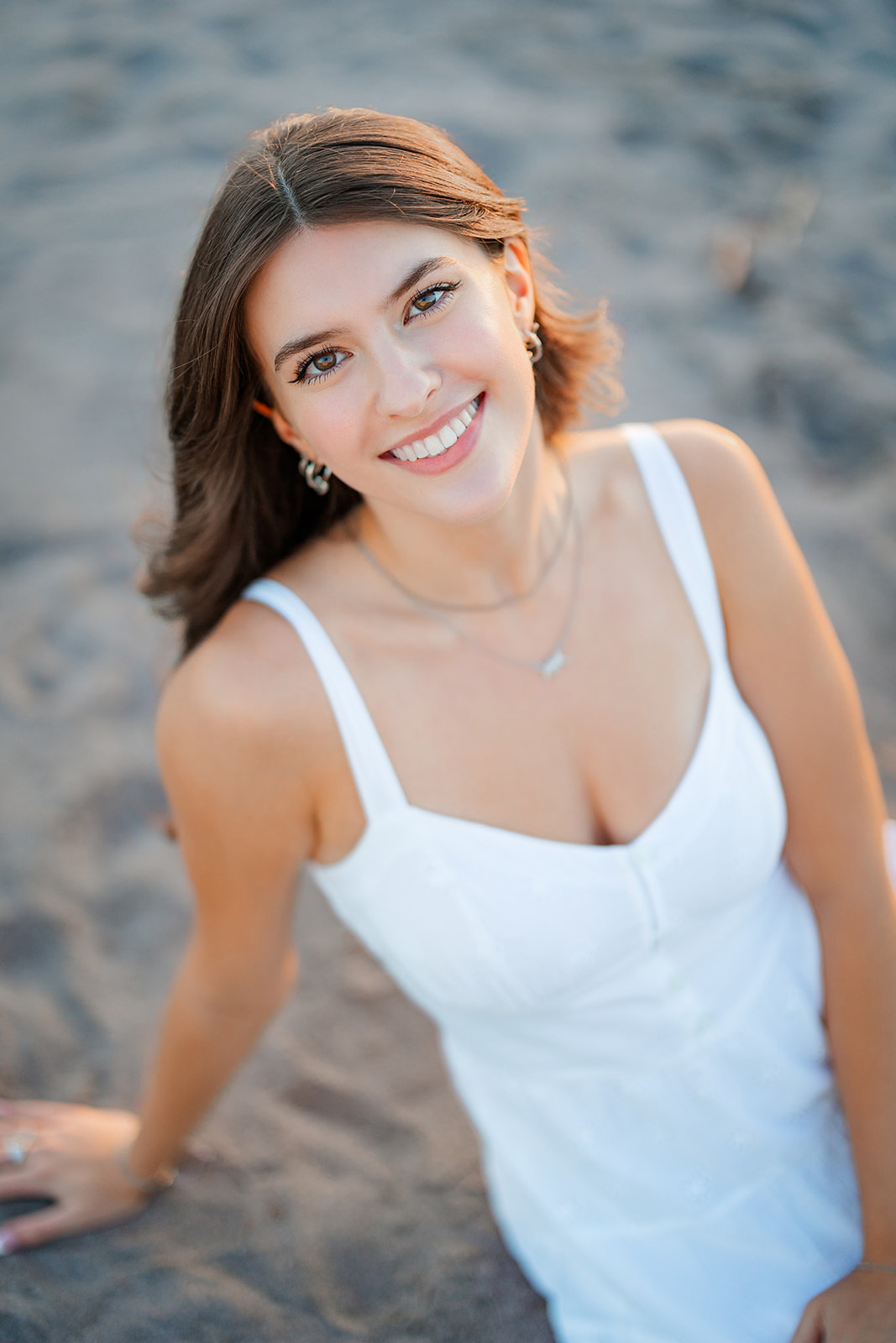 A teenage girl leans back onto her wrist while sitting on a beach at sunset in a white sun dress before finding her Prom Dress Bellevue