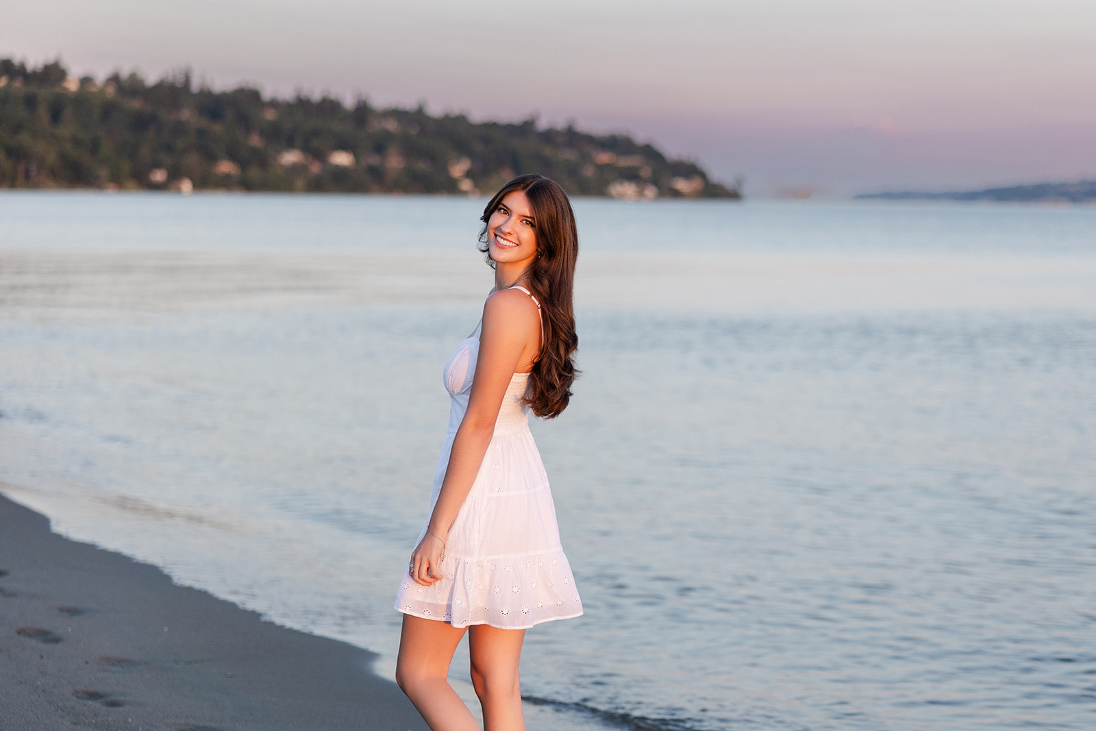 A teenage girl in a white dress walks on a beach at sunset while smiling over her shoulder