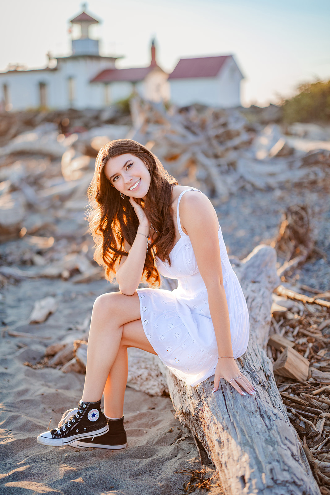 A teen girl sits on a driftwood log on a beach at sunset in a white dress and converse after visiting Seattle Boutiques