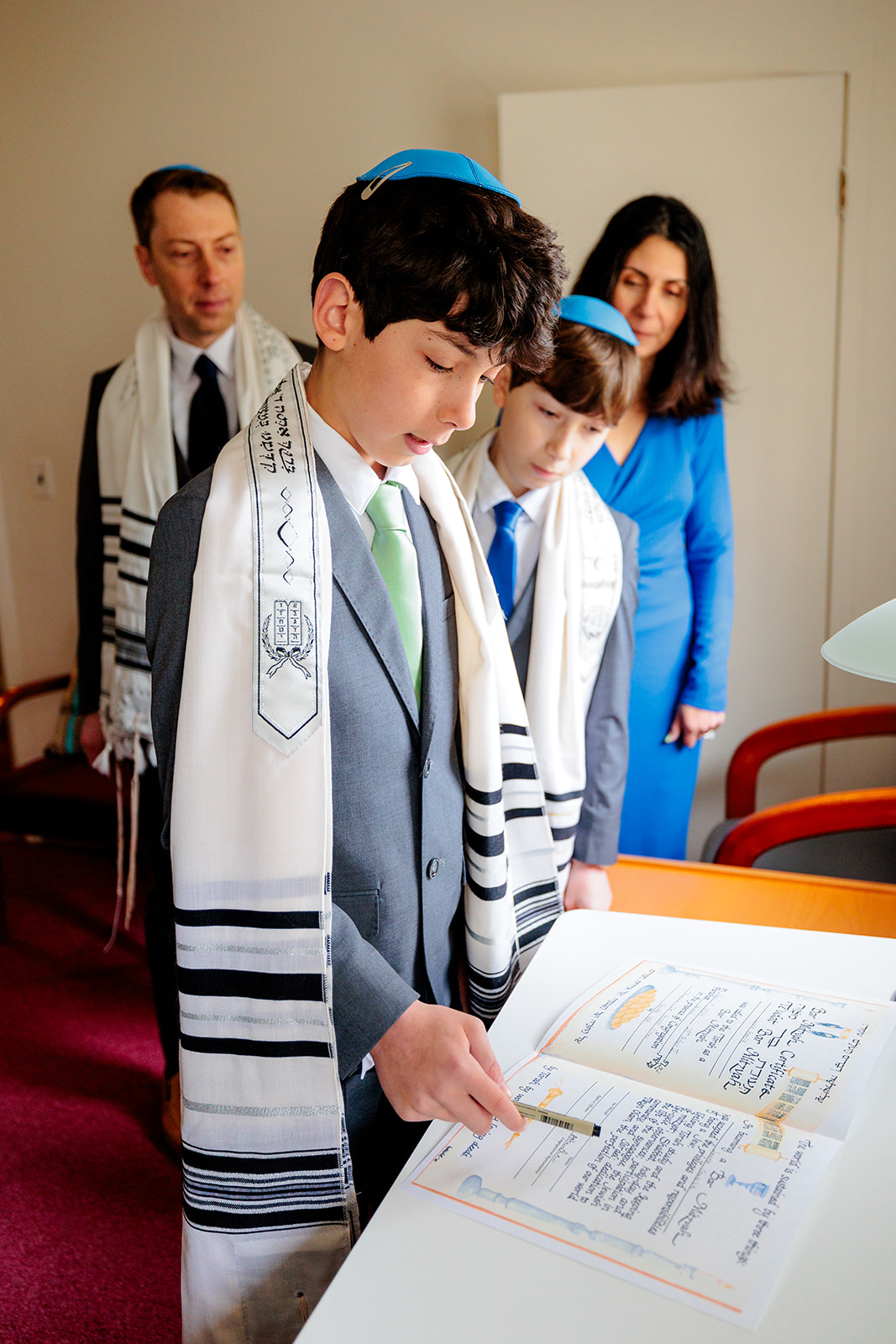 A teen boy reads from the torah with his Rabbi and mom looking on