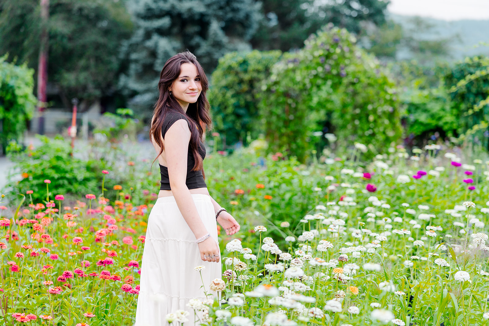 A high school senior walks through a colorful garden in a black shirt and white skirt while smiling over her shoulder after visiting Florists In Seattle