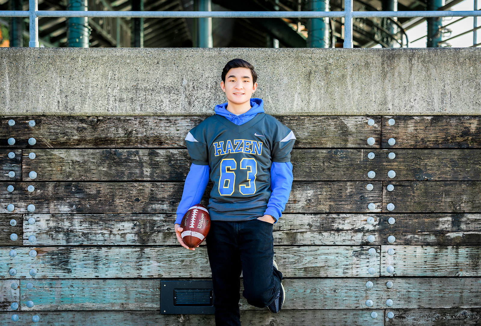 A Hazen High School senior football player leans against a stadium wall in his jersey while holding a football