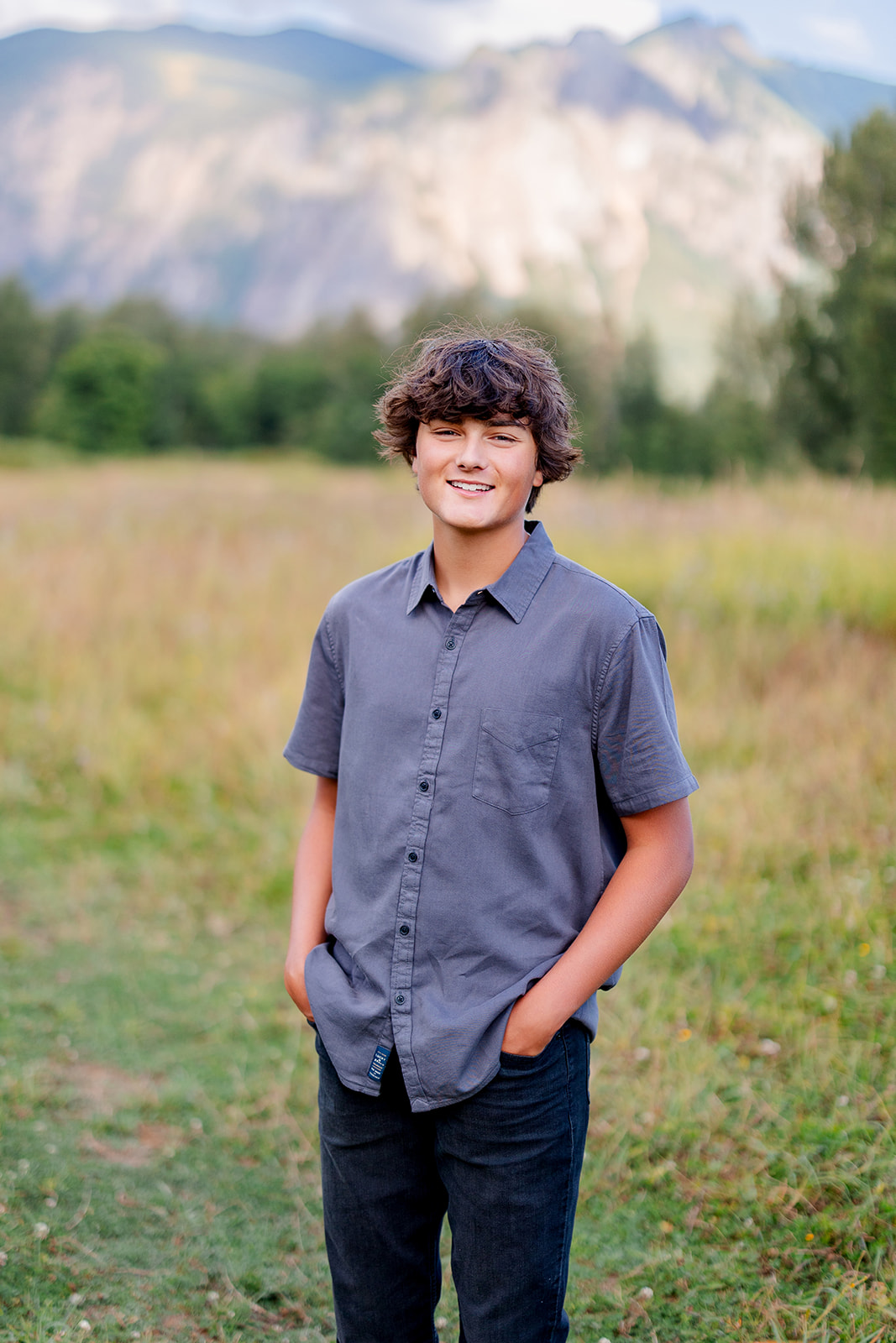 A smiling High School senior in a grey shirt stands in a field by the mountains with hands in his pockets