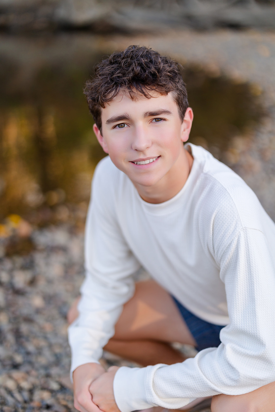 A Hazen High School senior squats near a river in a white long sleeve shirt