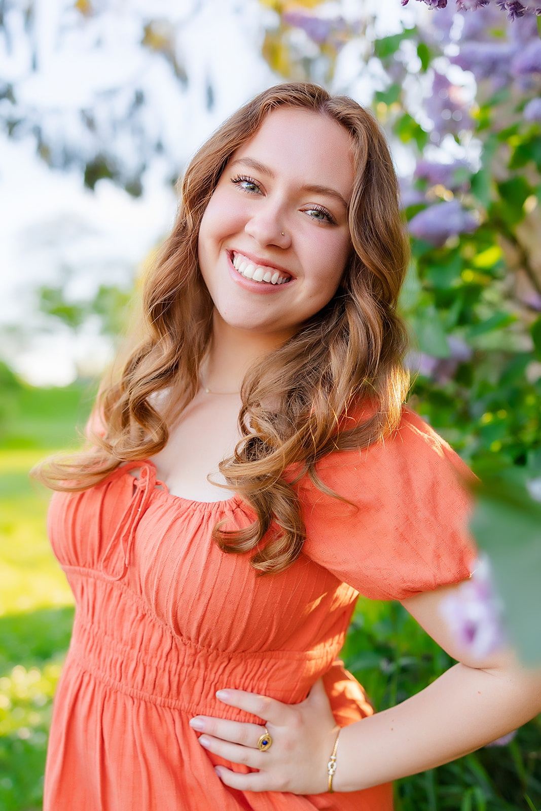 A happy high school senior leans on her hip in an orange dress under a blooming bush after shopping for Homecoming Dresses Bellevue