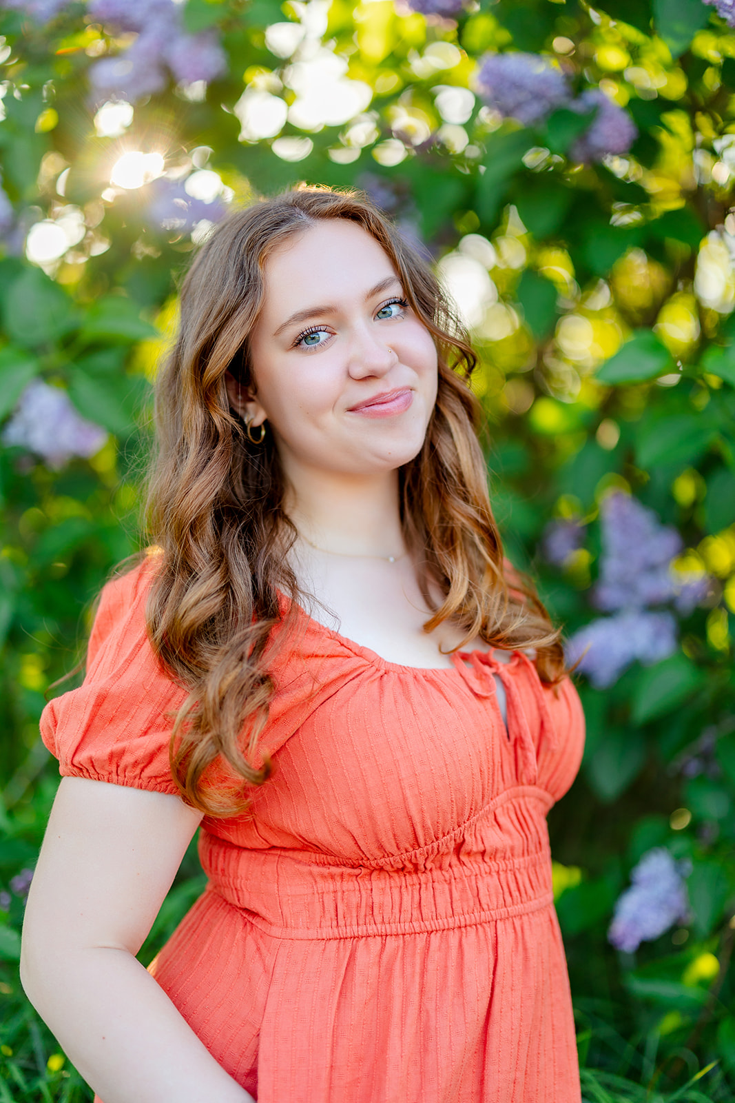 A high schools senior in an orange dress stands smiling in a garden after shopping for Homecoming Dresses Bellevue