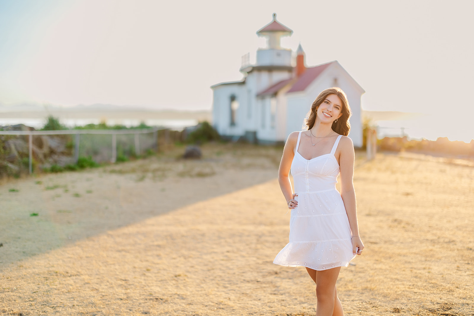 A woman in a white dress stands by a lighthouse at sunset with a hand on her hip after shopping for Homecoming Dresses Seattle