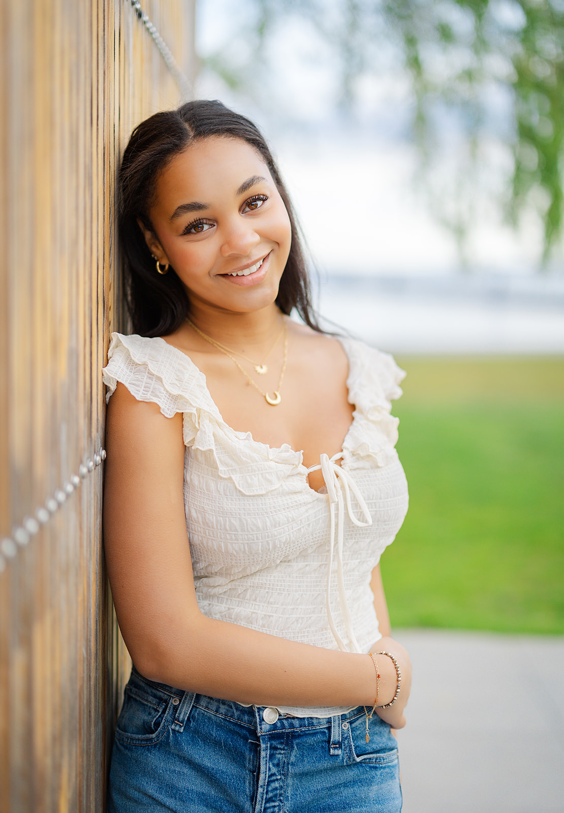 A senior in high school leans on a wooden park fence smiling in a white top and jeans