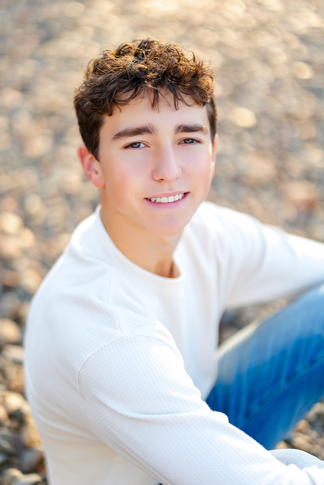 A young man sits on a rocky beach at sunset smiling in a white shirt