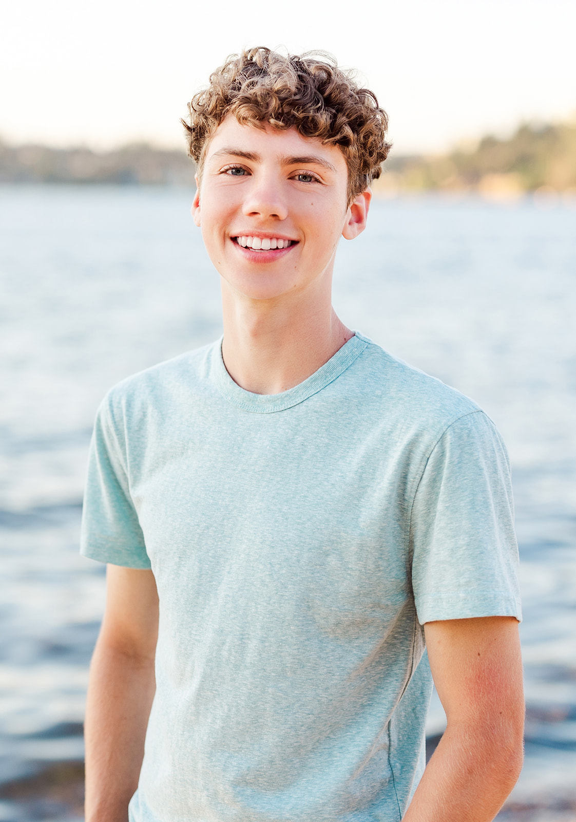 A Kirkland Private Schools graduate stands by the water while smiling in a green shirt at sunset