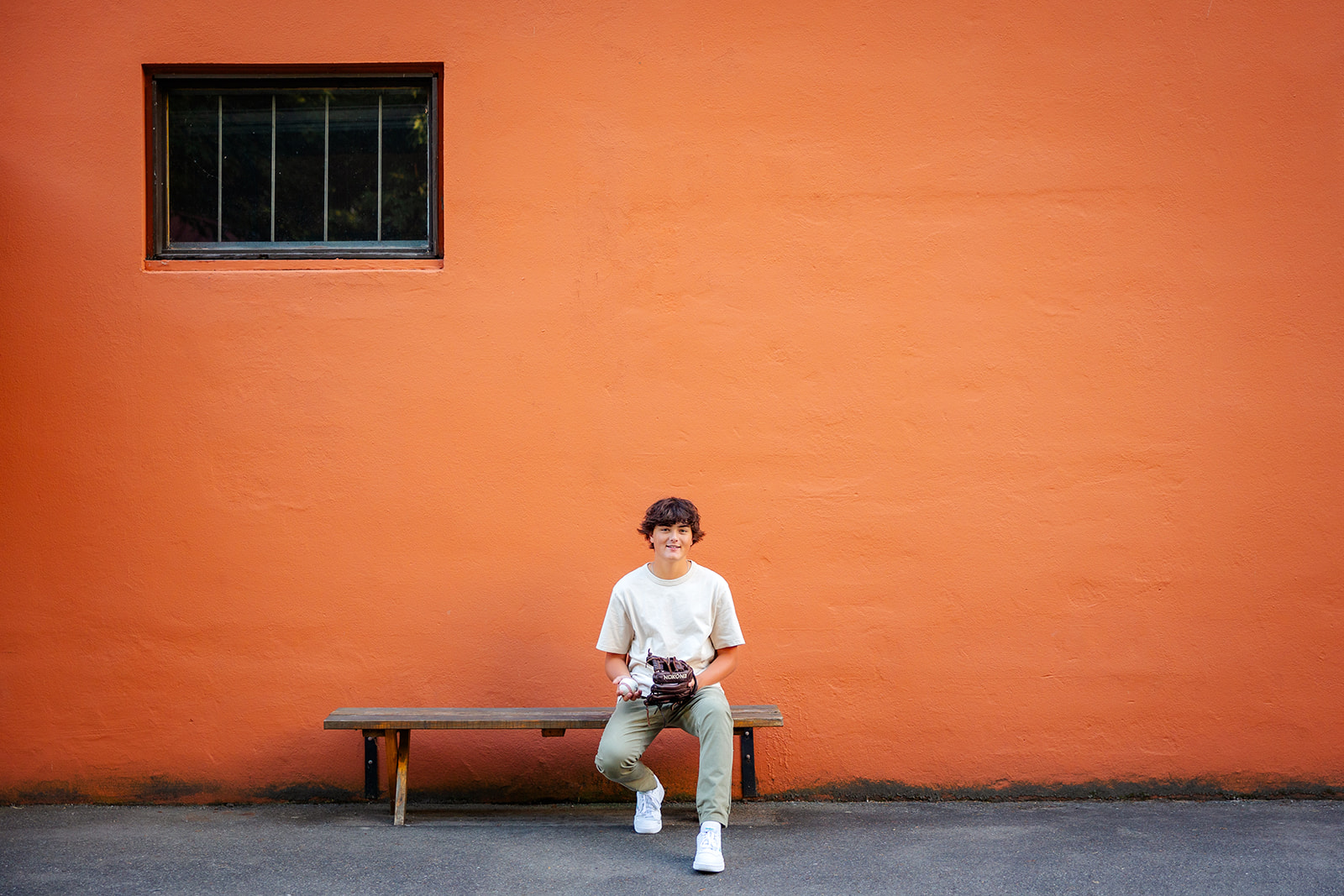 A boy sits on a bench by an orange wall with a glove and baseball at Kirkland Private Schools