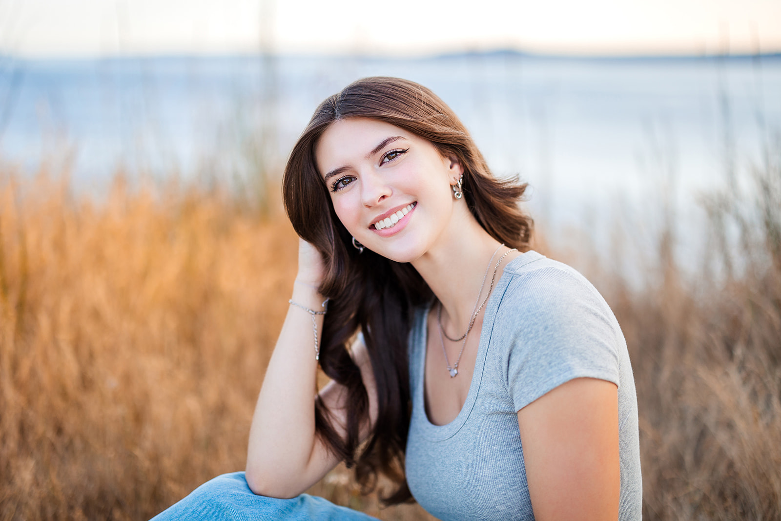 A senior in high school smiles while sitting near the water with a hand in her hair