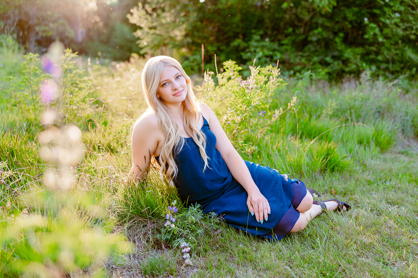 A high school senior sits in a park trail with wildflowers at sunset in a blue dress