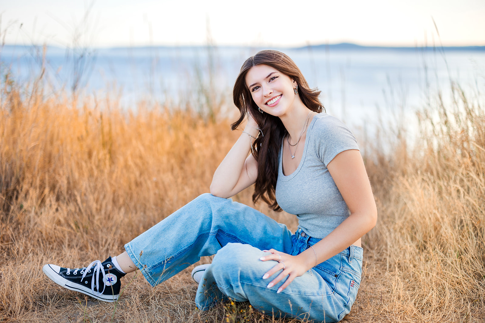 A teenage girl with dark hair sits by the water in tall golden grass leaning on her knee