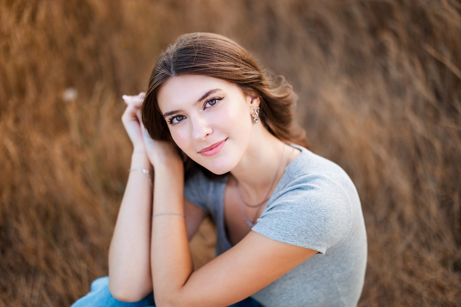 A high school senior in a grey shirt sits in a field of tall golden grass after visiting Nail Salons Renton, WA