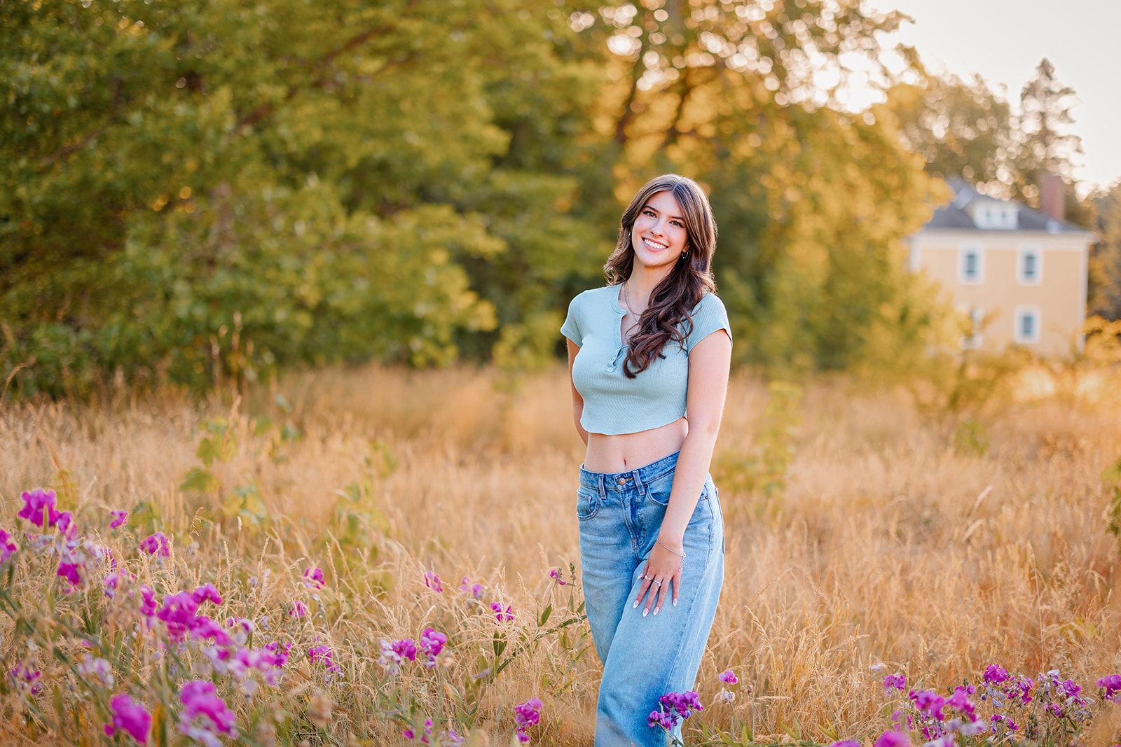 A high school senior smiles while standing in a field of golden grass at sunset