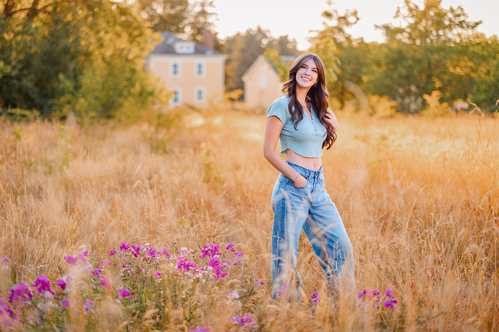 A woman in jeans and a short top stands in a field of golden grass and purple wildflowers at sunset after attending Newport High School