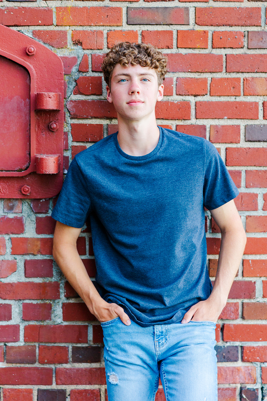 A High School senior leans on a brick wall in a blue shirt with hands in his jean pockets