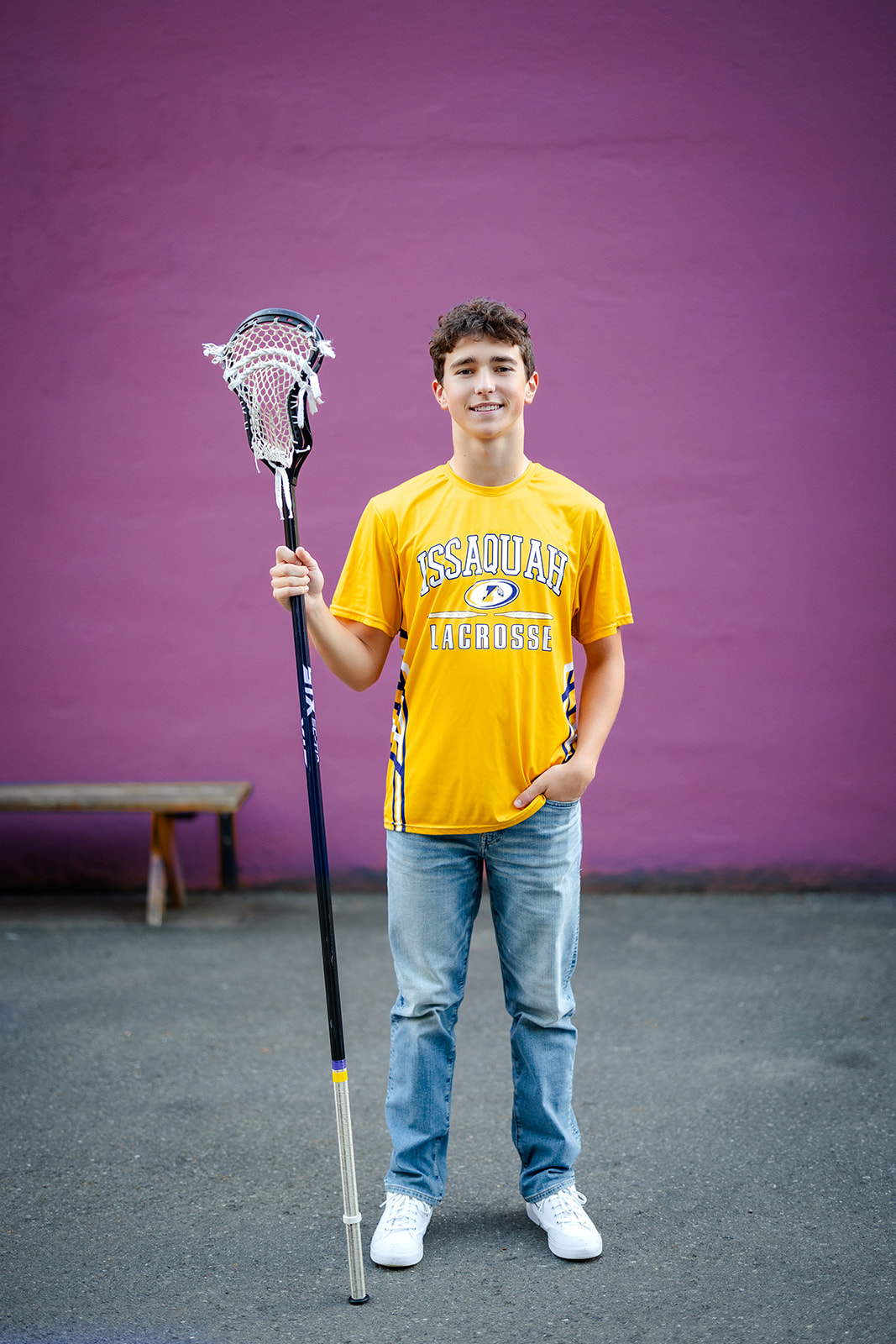 A Issaquah lacrosse player stands with his stick in front of a purple wall with a hand in his jeans pocket